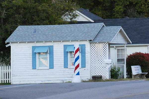Front view of the small  white wood-sided barber shop.  There is a large barber pole to the left of the building.